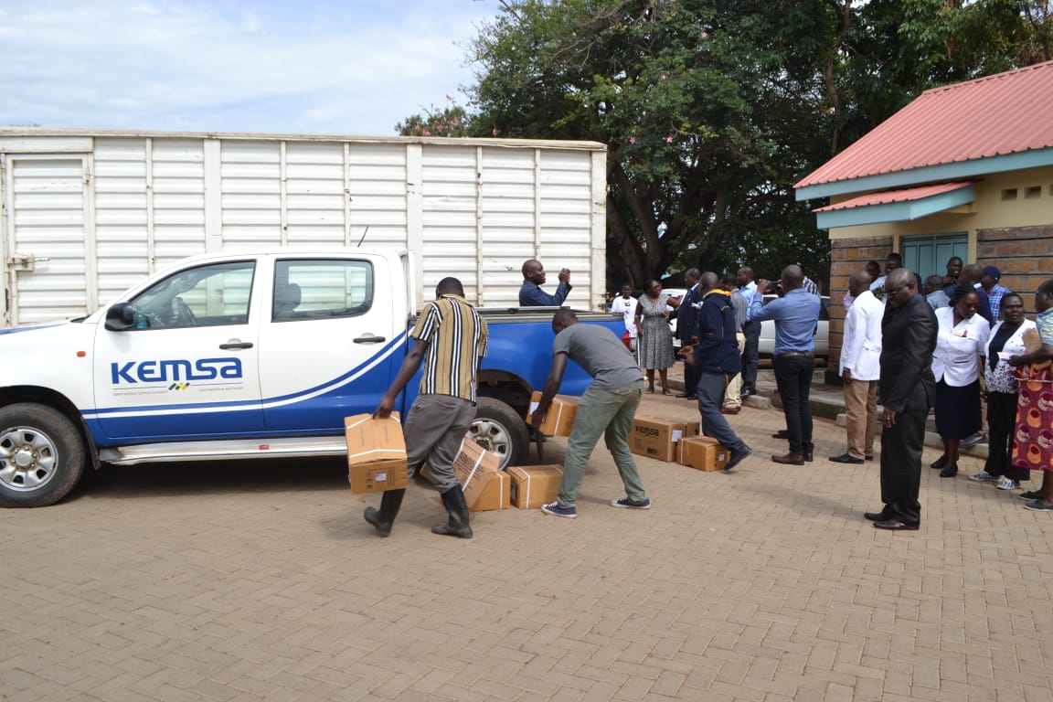 Offloaders carry the delivered medical commodities into the health facility as members of the public and the county Government watch.